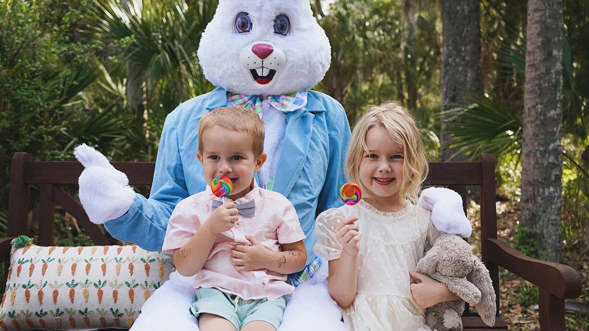 children holding lollipops posing for a photo with the Easter Bunny while sitting on a bench with greenery in the background