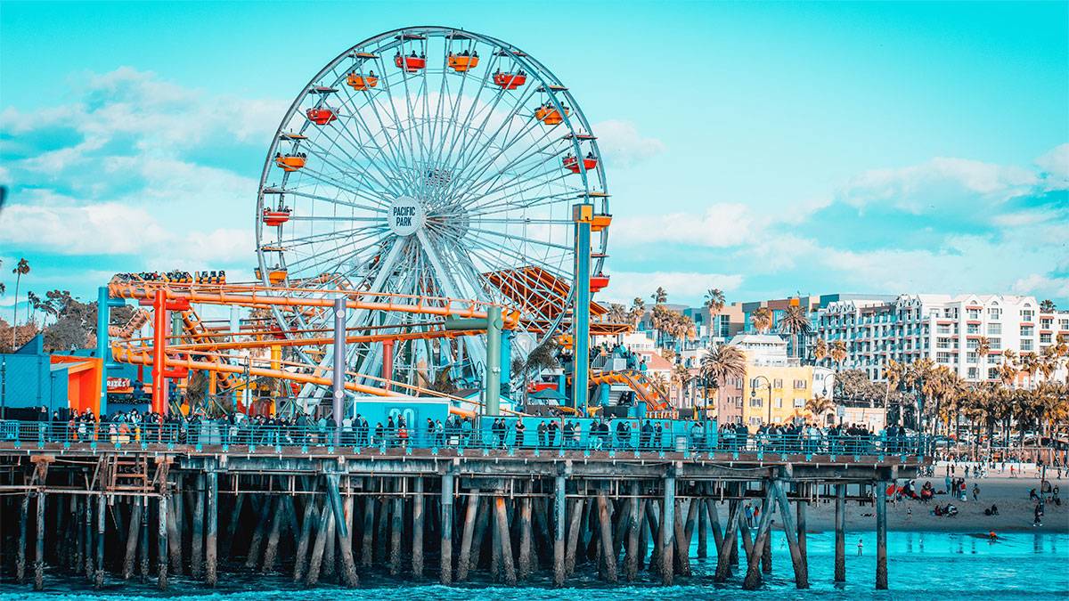 aerial view of santa monica pier with people, ferris wheel and other rides and view of shore and water and buildings and palm trees in background
