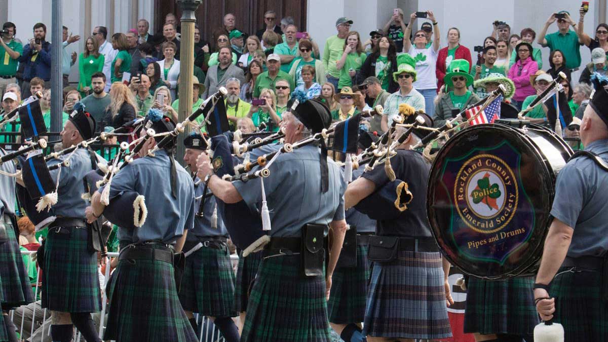 Close up of men marching in green kilts and grey shirts playing the bagpipes and drums with a crowd of people wearing green in the background at the Savannah St. Patrick's Day Parade in Savannah, Georgia, USA