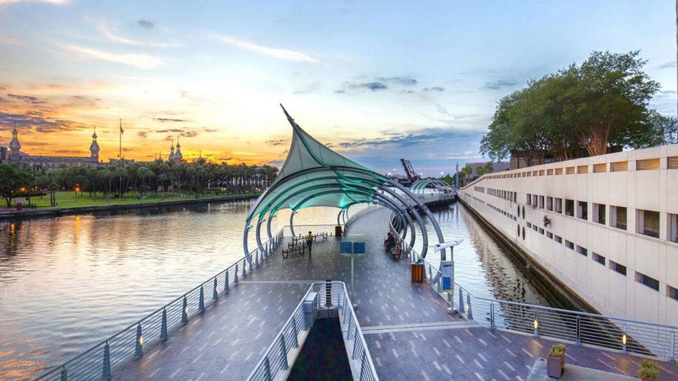 walk way over river with sculptural arch with trees and building in the distance at Tampa Riverwalk in Tampa Bay, Florida, USA