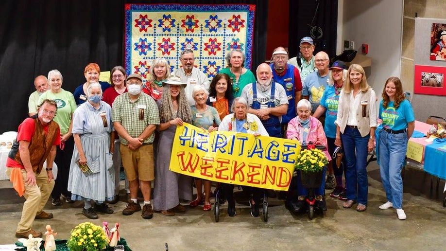 A group of older people with a quilt hanging behind them and four women in the front hold a sign that says Heritage Weekend at Southern Highland Craft Guild, Folk Art Center in Asheville, North Carolina, USA