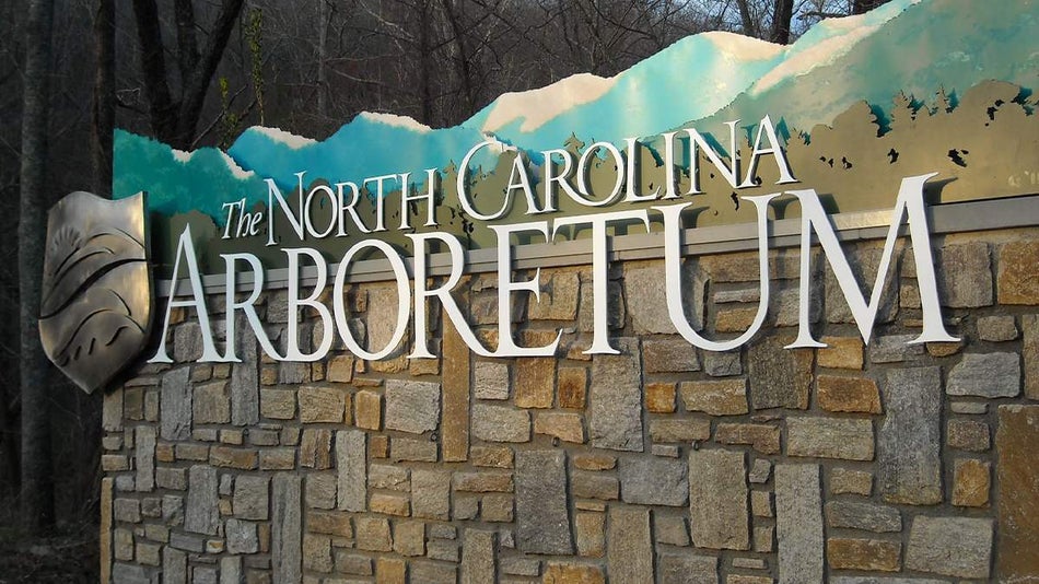 Close up photo of the sign for The North Carolina Arboretum, the bottom is stone and the top has different metal cut outs of trees and mountains in Asheville, North Carolina, USA