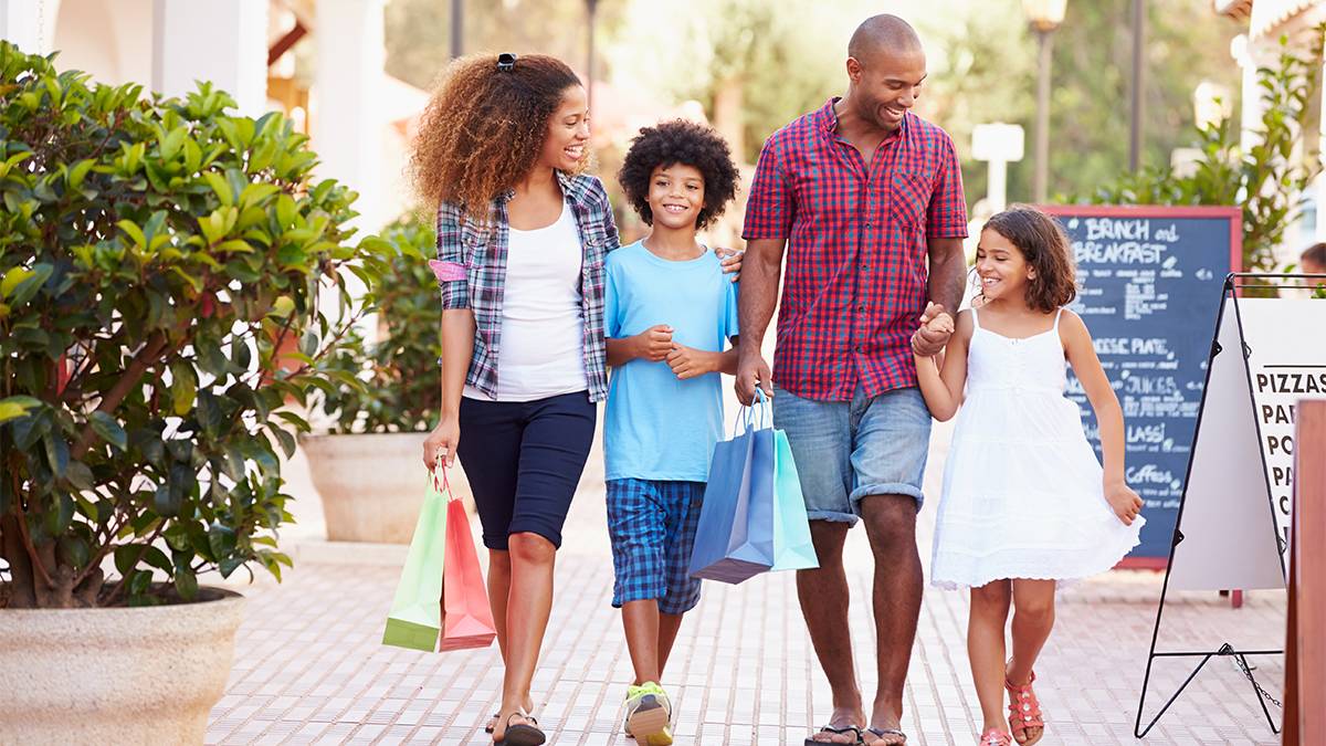 A family, two parents and two kids, holding hands and bags walking through an outdoor shopping area