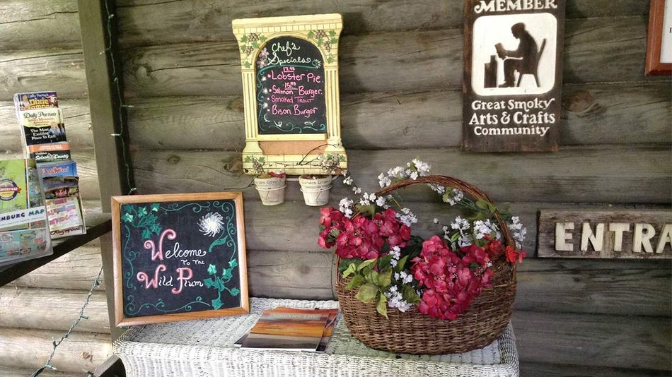 View of the entrance to Wild Plum Tea Room, a small white wicker tables with a welcome sign and a basket of flowers and a log wall behind it with a chef's specials menu on it in Gatlinburg, Tennessee, USA