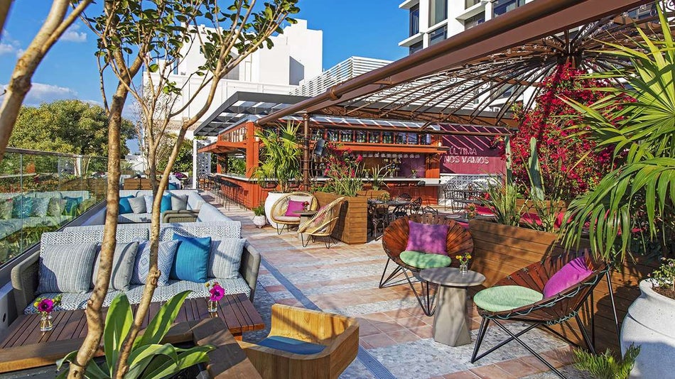 View of a colorful rooftop bar with lots of plants and different types of lounge seating with the bar towards the back and the city in the background at the Serena Rooftop Restaurant at the Moxy in Miami, Florida, USA