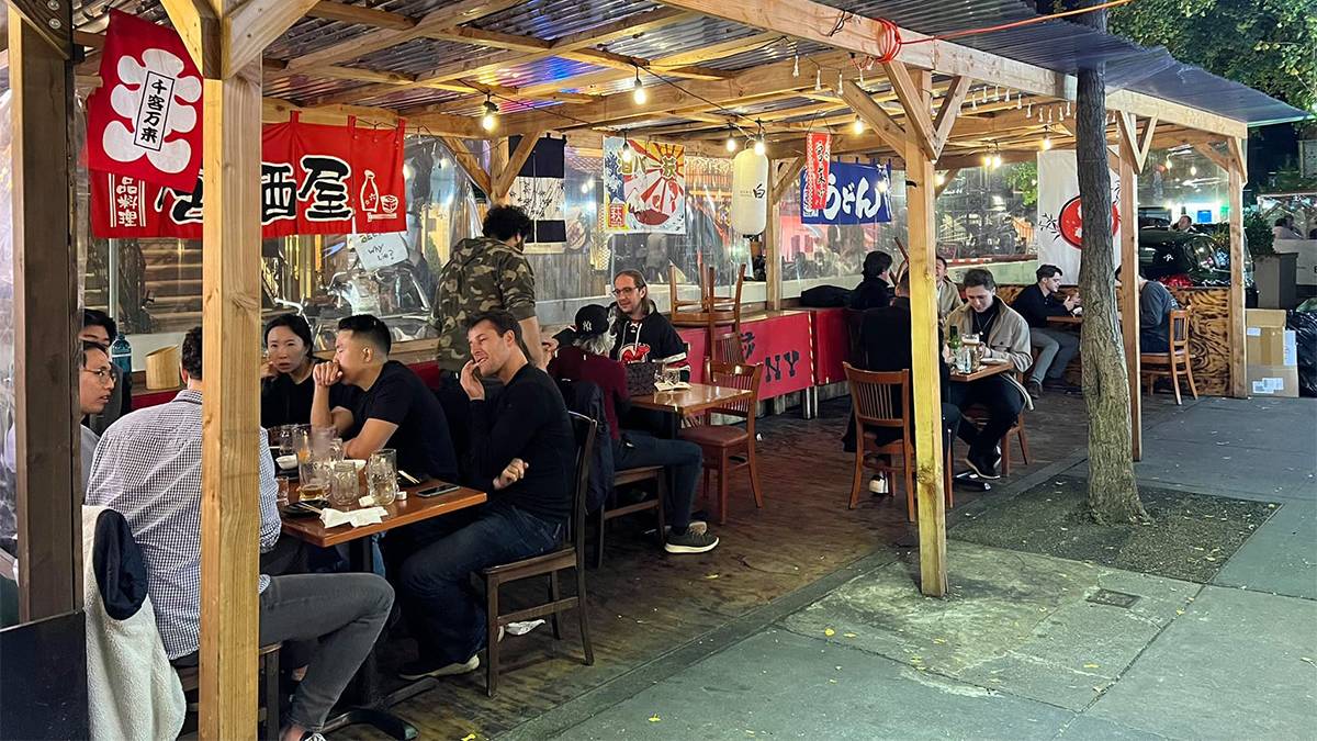 Close up of the outdoor dining area at Sake Bar Hagi full of people, a wooden structure open on one side and lots of string lights and posters on the tin walls in NYC, New York, USA