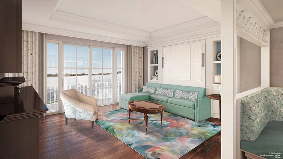 View of a living room area with a large window doors that lead to a balcony and a light blue couch, brown coffee table, and a cream colored chair with white walls and brown shelving on the left at the Disney Vacation Club Resort Studios in Orlando, Florida, USA