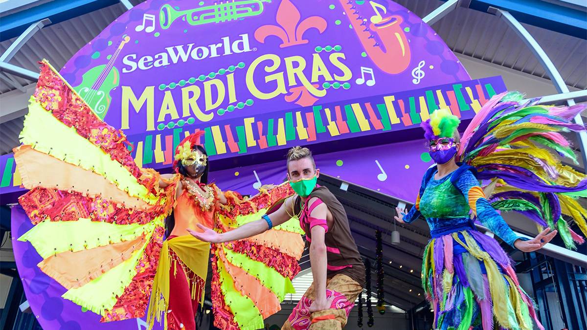 Close up of three performers in bright colorful Mardi Gras costumes with a large purple sign behind them for SeaWorld Mardi Gras in Orlando, Florida, USA