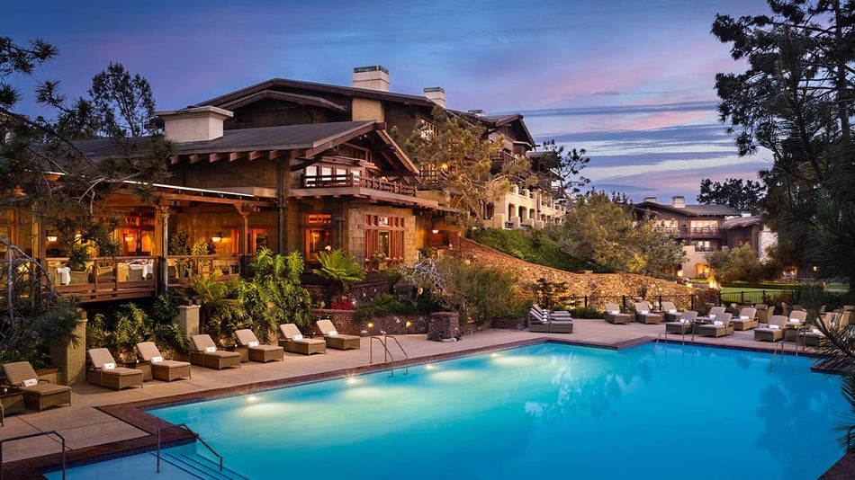 View of the Torrey Pine Lodge, a large multi level wooden and stone building with an outdoor dining area and it's pool lounge area with a light purple sunset in the background in San Diego, California, USA