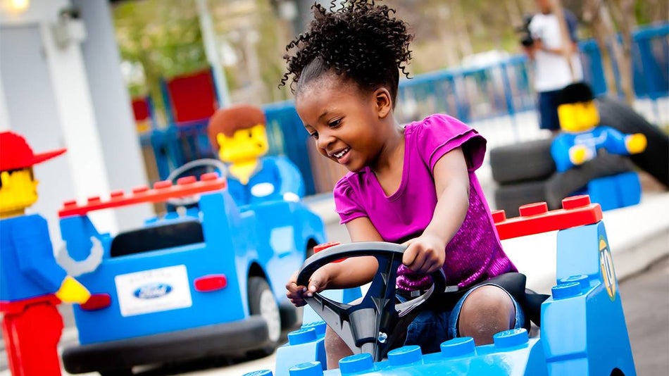 Close up photo of a little girl in a pink shirt driving a blue LEGO car with LEGO people in the background at driving school LEGOLAND in San Diego, California, USA