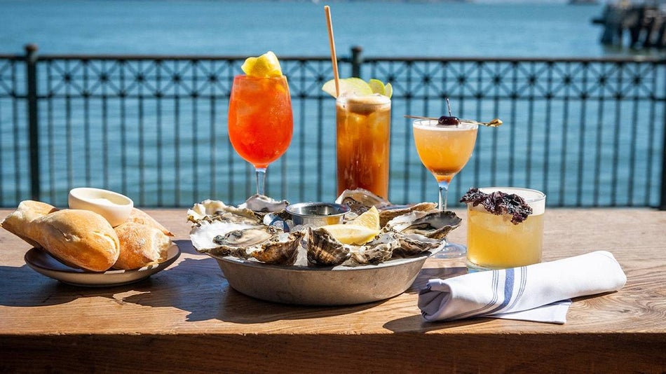Close up of a plate of oysters with a side of bread and four different cocktails on a wooden table with the ocean behind them at Hogs Island Oyster Company in San Francisco, California, USA