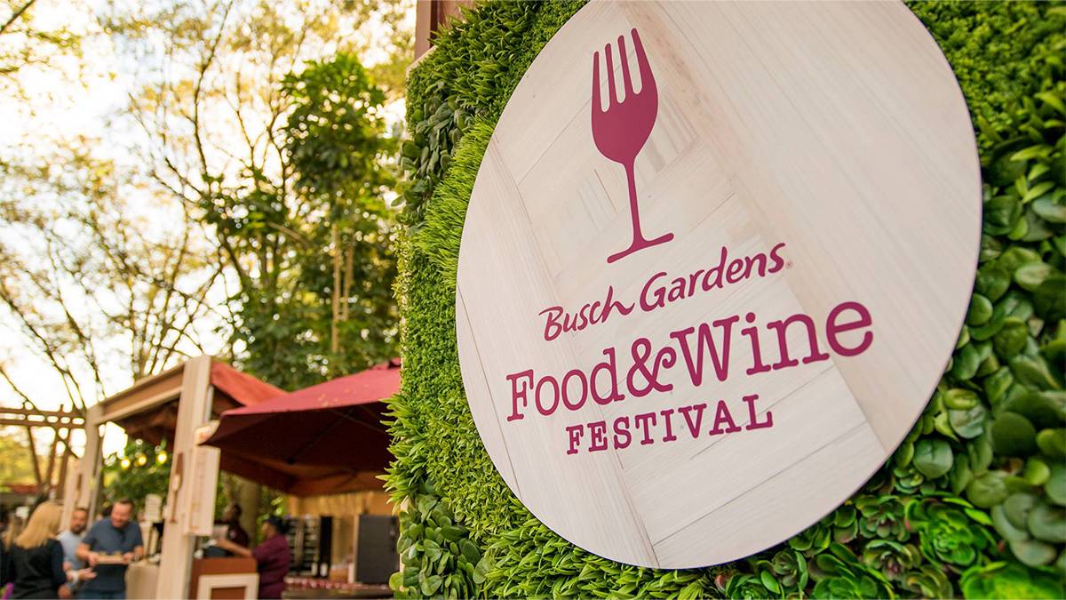 Close up of a circular wooden sign with purple lettering for the Busch Gardens Food and Wine Festival on a shrub with people in the background in Tampa, Florida, USA