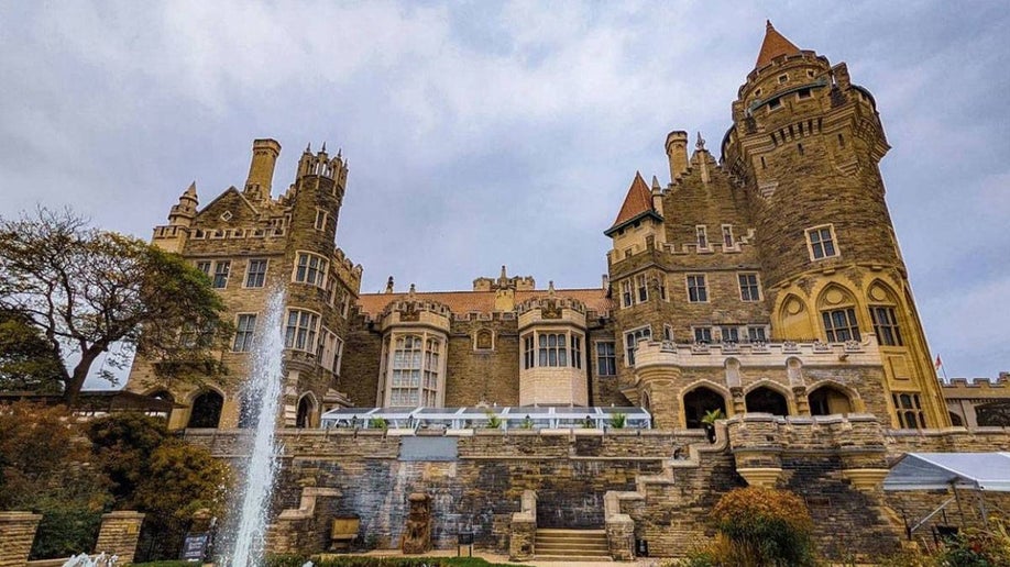 View looking up at Casa Loma Castle, a large castle made out of brown and grey stone, with white clouds behind it in Toronto, Ontario, Canada