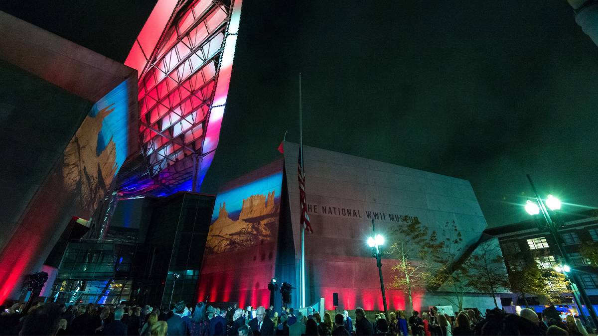 View looking up at the National WWII Museum at night with red, white, and blue lights and a crowd of people below it